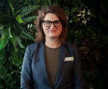 Woman (Kate Eastoe) wearing corporate attire and glasses, smiling. She has a leafy background.