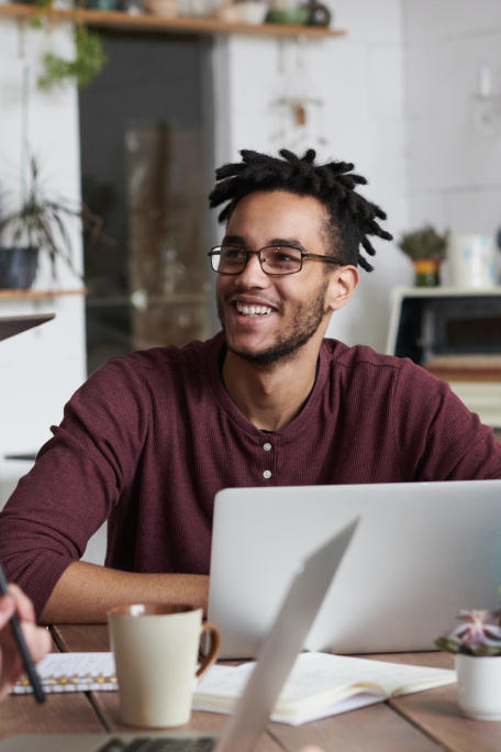 Man with dreadlocks and glasses sitting in front of laptop. Smiling off to the side.