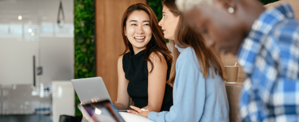 Woman smiling to another woman, whilst third woman out of focus works on laptop