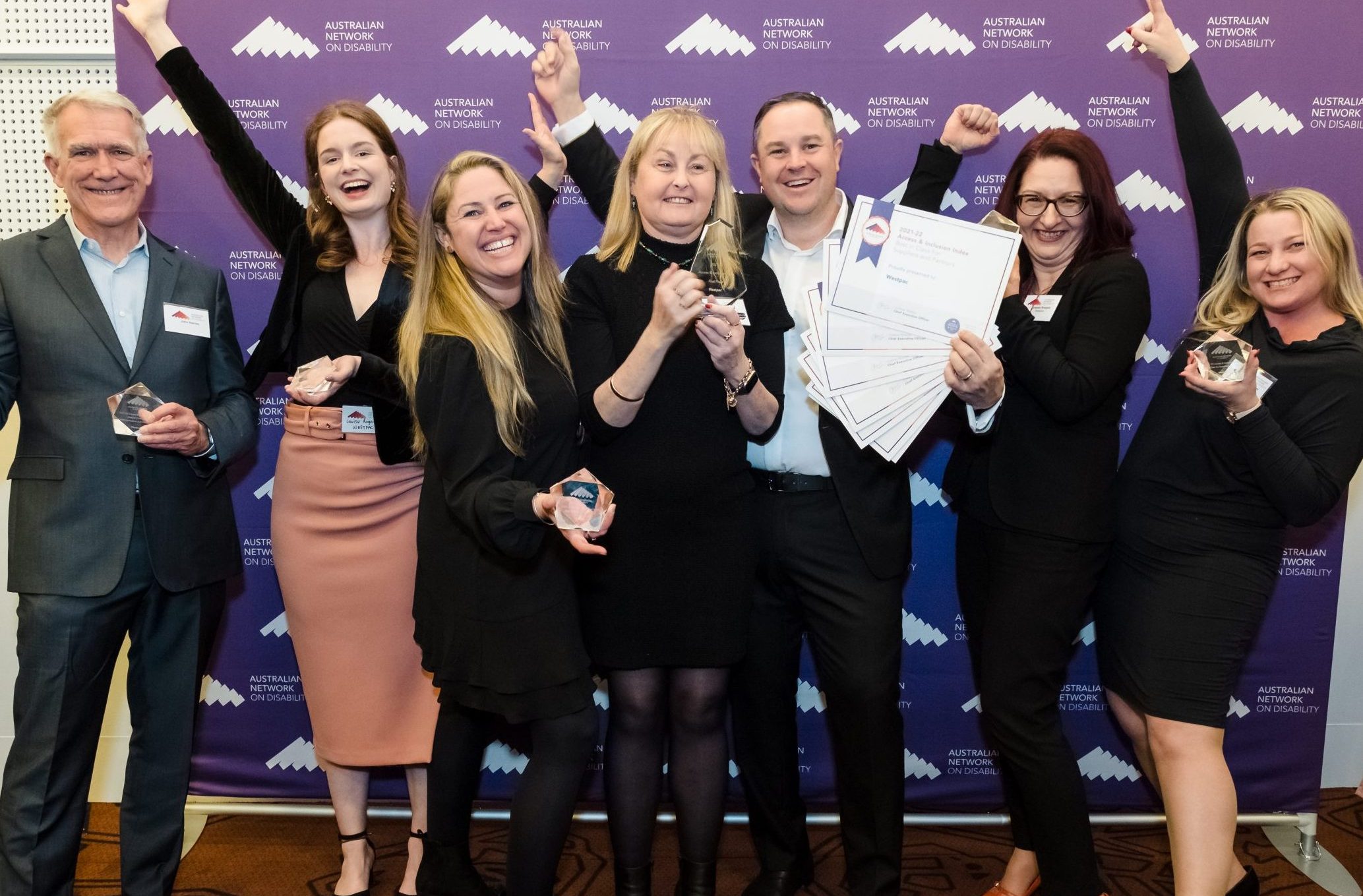 Group of seven people standing holding certificates and awards, smiling, with their hands in the air in celebration. Purple Australian Network on Disability banner behind them.