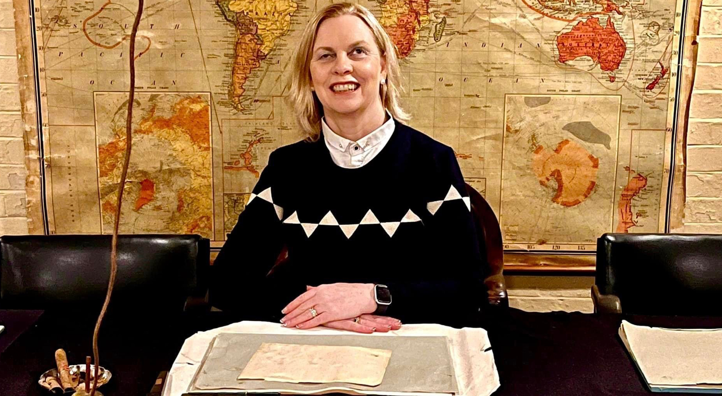 Donna, a woman with short blonde hair, is wearing a collared shirt and black jumper with white rectangles. Her hands are folded on her desk.