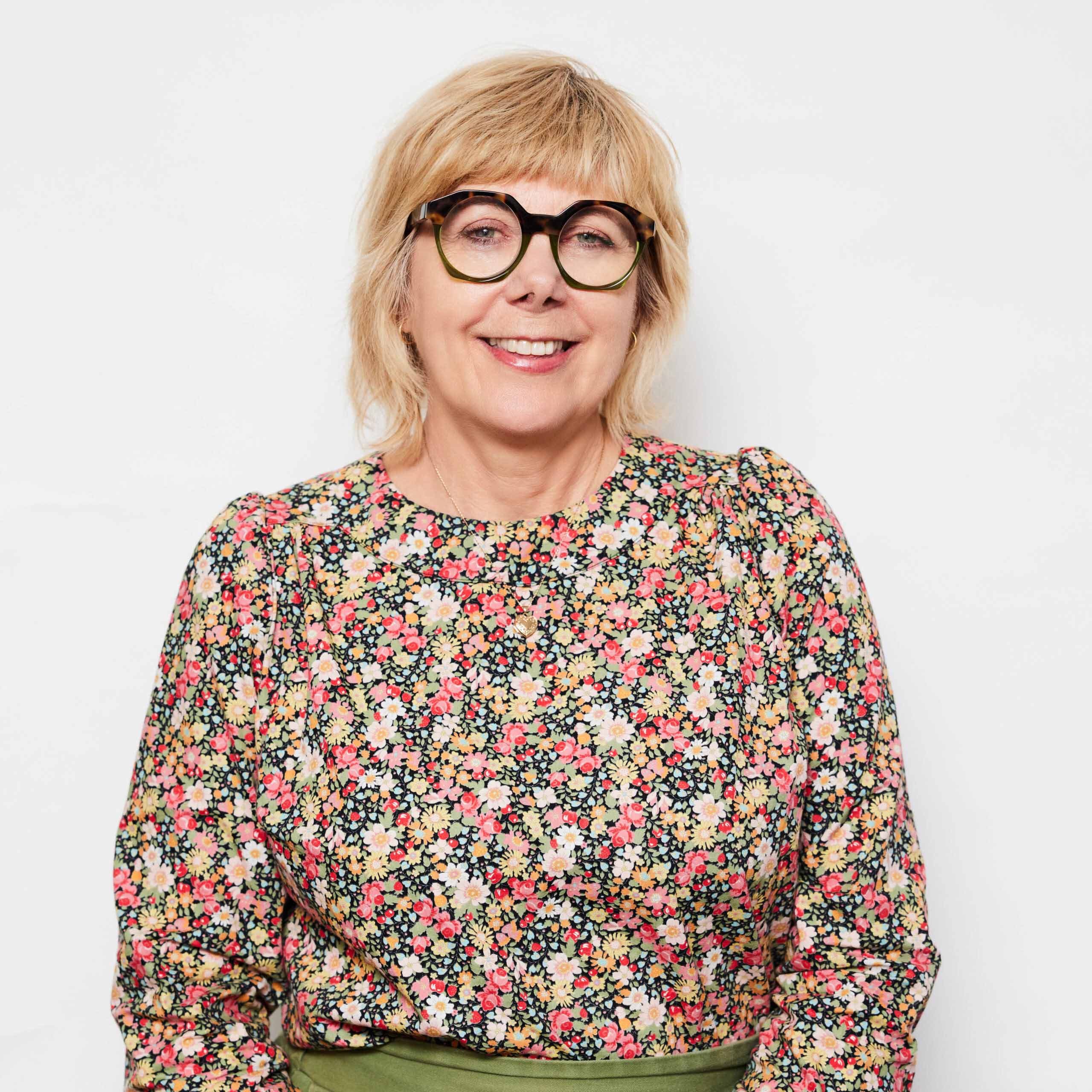 Penny, a woman with short hair and glasses. She is wearing a floral top. She is smiling.